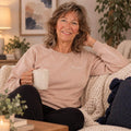 Woman in beige crewneck sweatshirt holding mug, smiling on cream couch in warm living room.