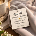 Cream tag attached to folded gray crewneck sweatshirt beside coffee mug and white flowers.