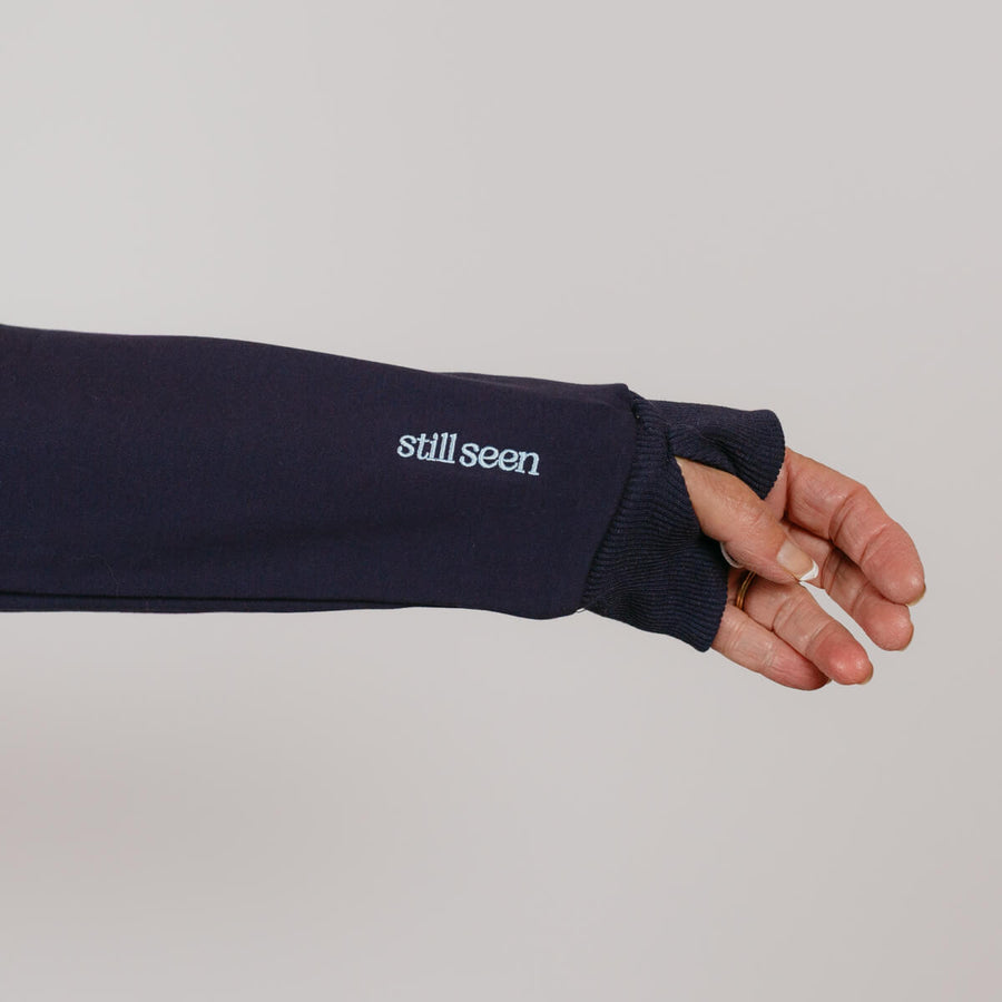 Navy crewneck sweatshirt with "still seen" text displayed on extended sleeve worn by person's arm.