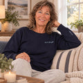 Woman in navy crewneck sweatshirt sitting on striped couch, smiling at camera in bright living room.