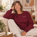 Woman with wavy shoulder-length hair smiling while sitting on a couch in a burgundy crewneck sweatshirt.