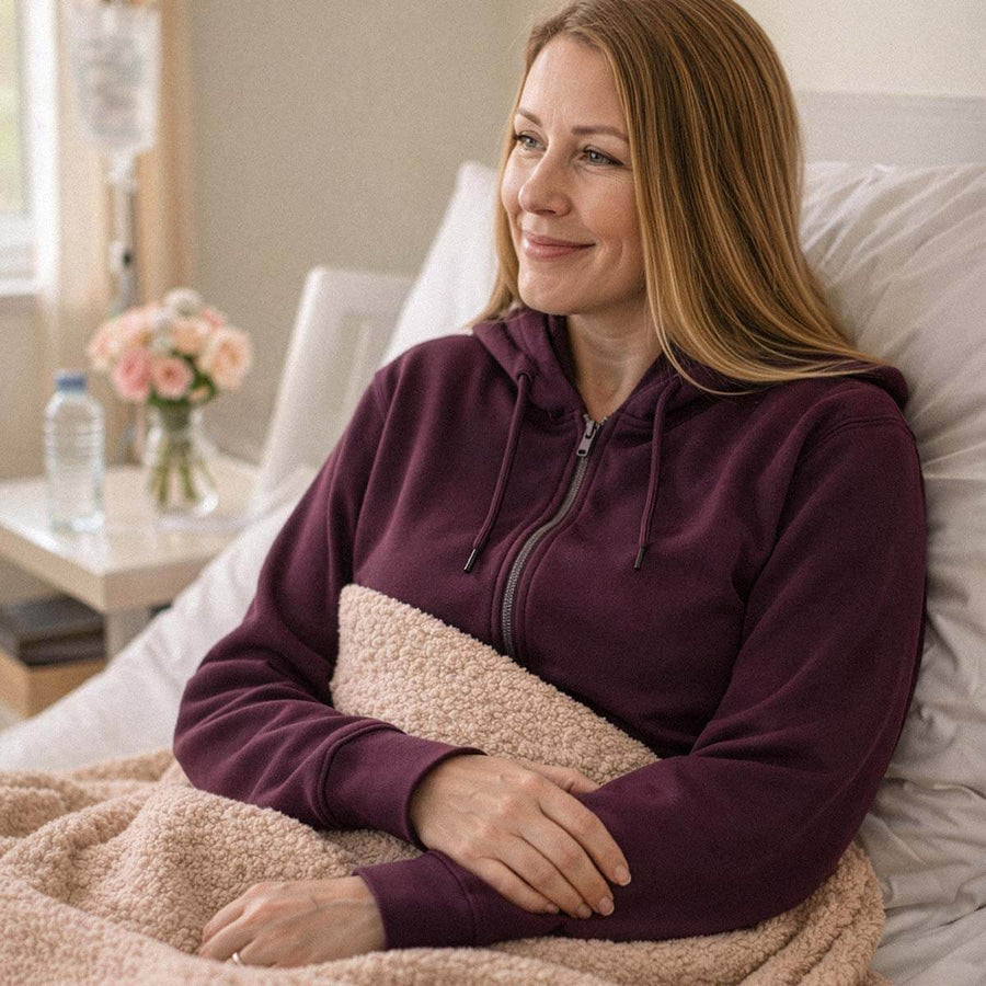 Woman in burgundy hoodie sitting on bed, wearing adaptive clothing, smiling toward window.