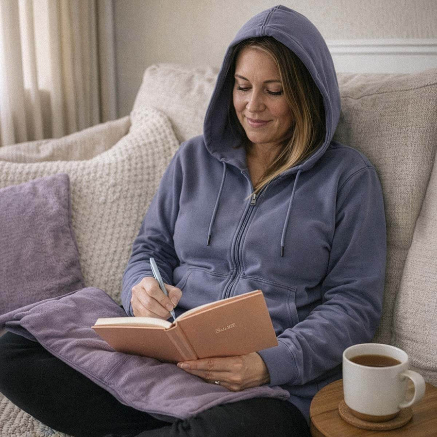 Woman in blue hoodie writing in notebook while seated on couch with coffee mug nearby.