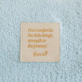 Close-up of cream infusion blanket with gold inspirational quote and BeWell branding detail.