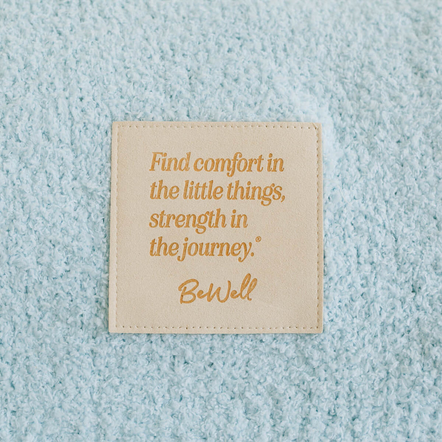 Close-up of cream infusion blanket with gold inspirational quote and BeWell branding detail.
