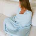 Woman wrapped in light blue infusion day blanket sitting on cream couch lifestyle shot.