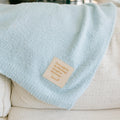 Light blue infusion day blanket draped over cream chair with tan product label visible.
