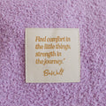 Close-up of cream infusion day blanket with gold inspirational quote detail on purple textured surface.