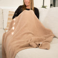 A woman sitting on a white couch under a tan fuzzy blanket.