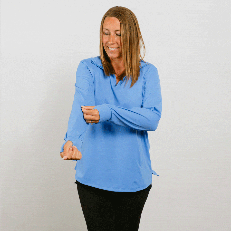 Woman wearing light blue quarter zip pullover and black pants smiles while gesturing with both hands.