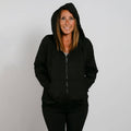 Woman wearing a black zip-up hoodie with hands in pockets, smiling at camera against white background.