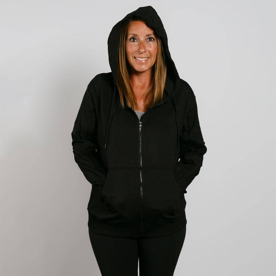 Woman wearing a black zip-up hoodie with hands in pockets, smiling at camera against white background.