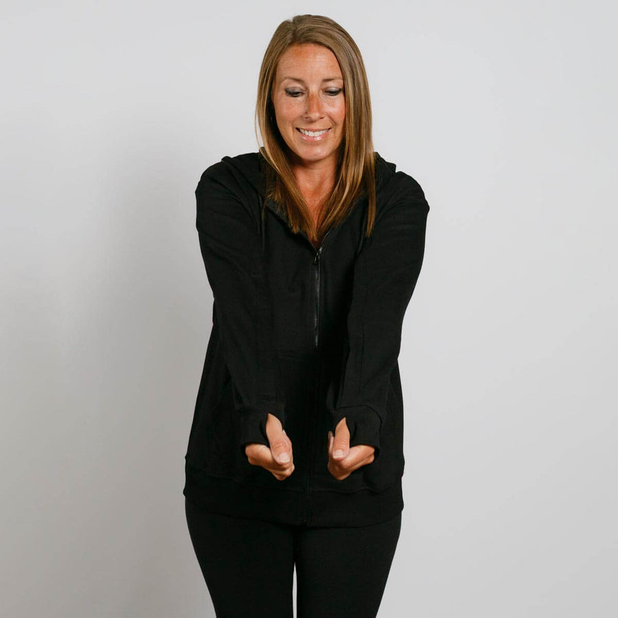 Woman wearing black zip-up hoodie with hands in front pockets, smiling downward against white background.