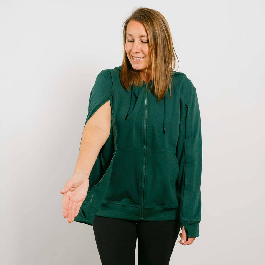 Woman wearing a dark green zip-up hoodie with black pants against a white background.