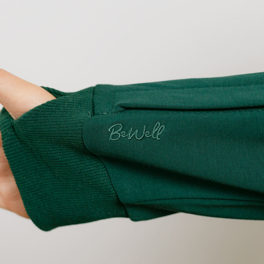 Person wearing a dark green zip hoodie with ribbed cuffs and embroidered text on the sleeve.