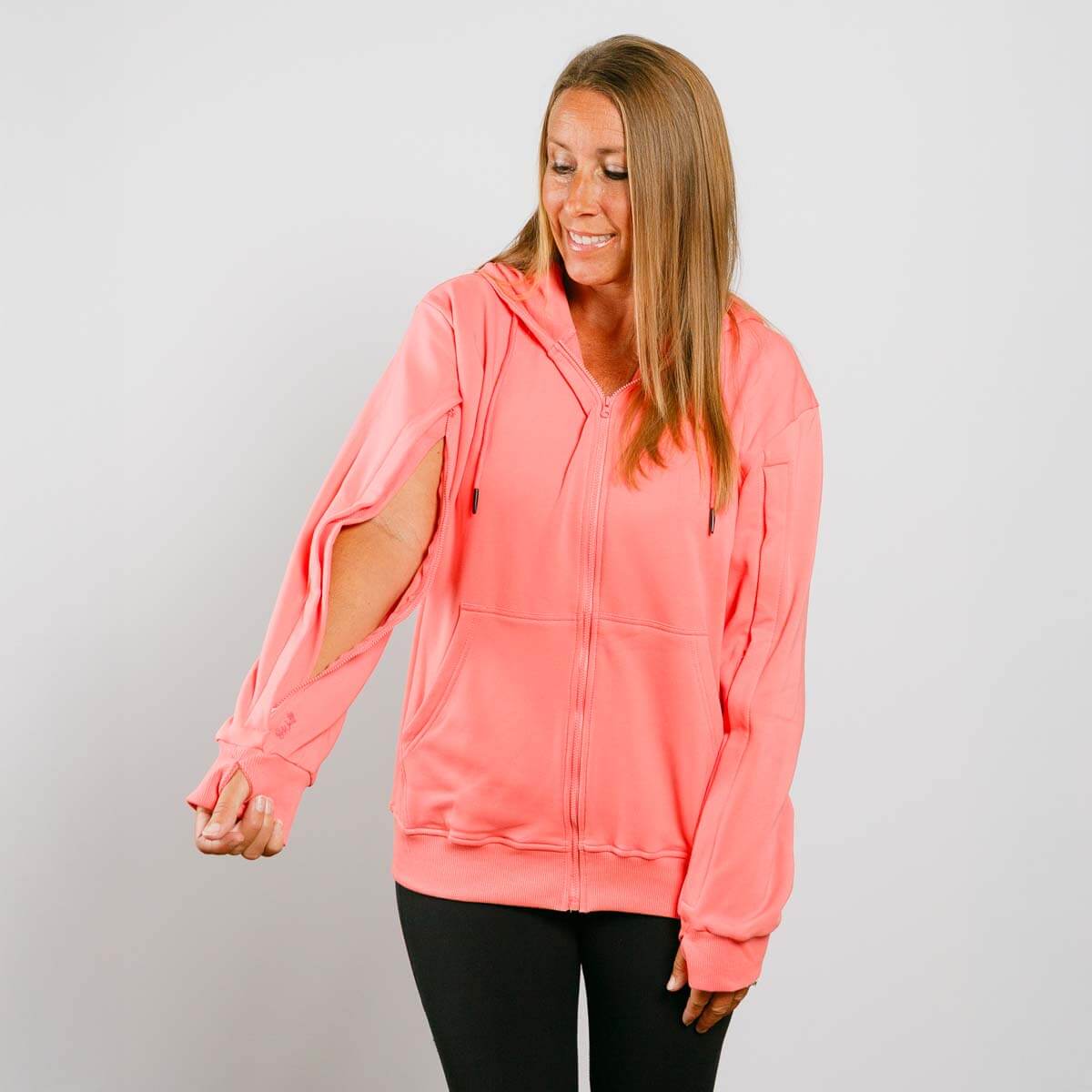 Woman wearing a coral pink hoodie with cut-out sleeves on a gray background   | Model is 5'3" 130lbs wearing size S