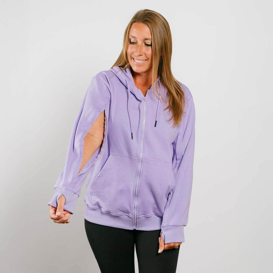 Woman wearing a lavender zip-up hoodie with thumbholes and black pants, smiling downward.