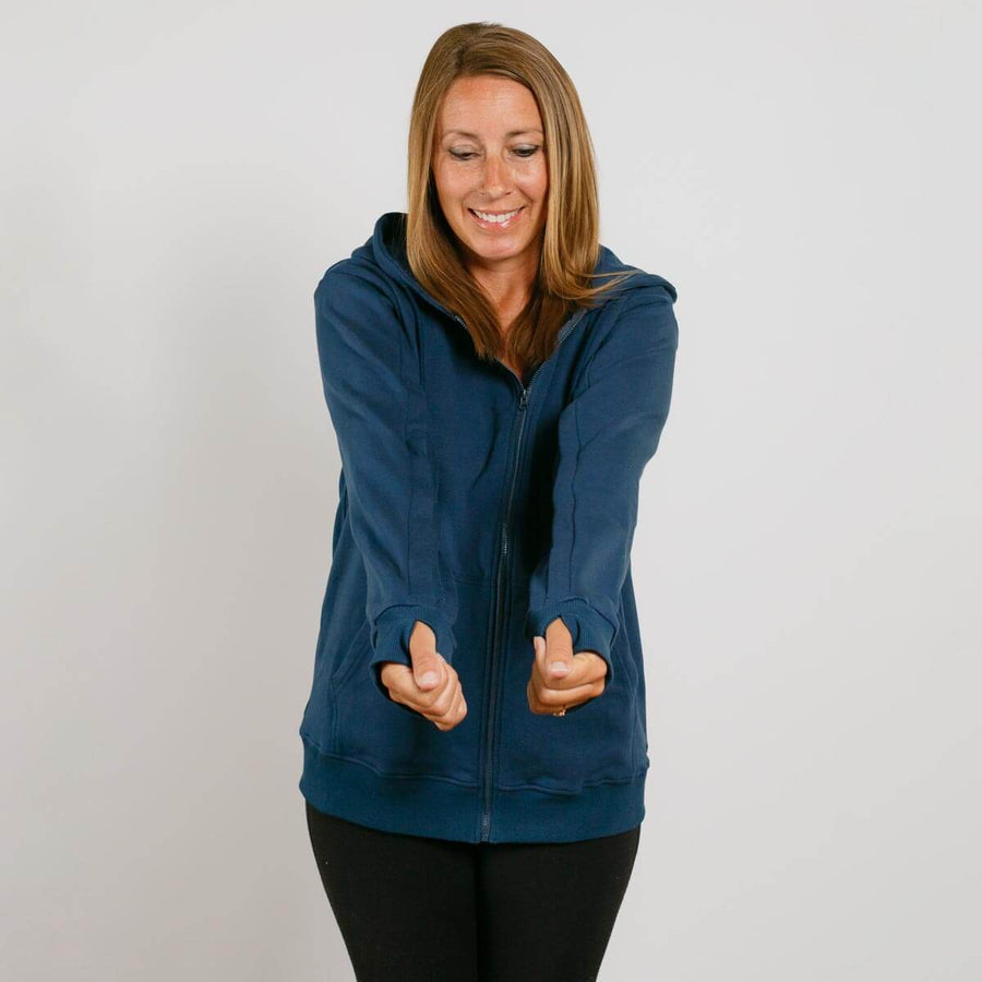 Woman wearing a navy blue zip-up hoodie with black pants, smiling with hands gesturing outward.