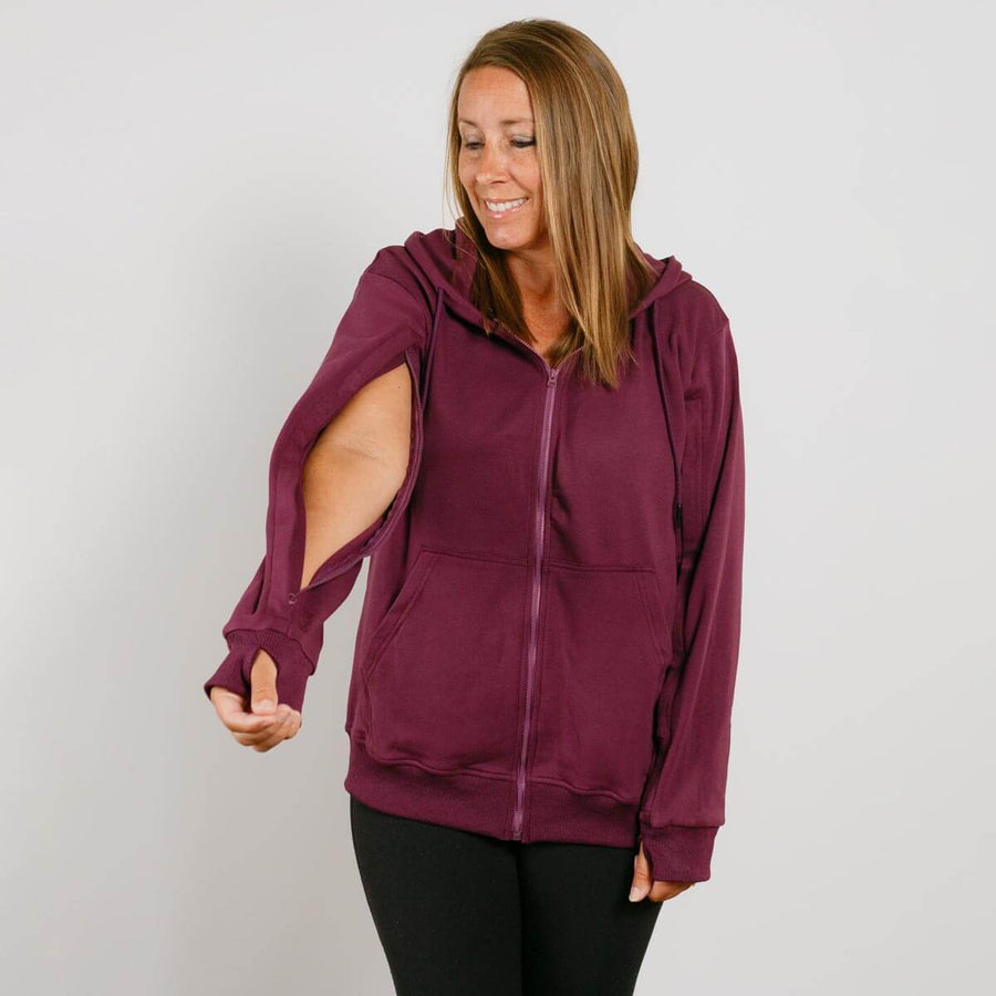 Woman wearing a burgundy zip hoodie with cutout sleeves over black leggings, smiling downward.