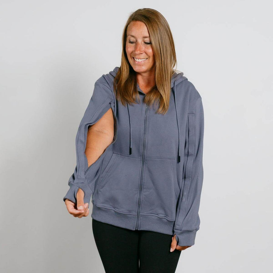Woman wearing a slate blue zip-up hoodie with thumb holes and black pants, smiling with eyes downcast.