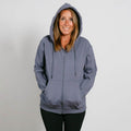 Woman wearing a slate blue zip-up hoodie with hands in pockets, smiling at camera against white background.