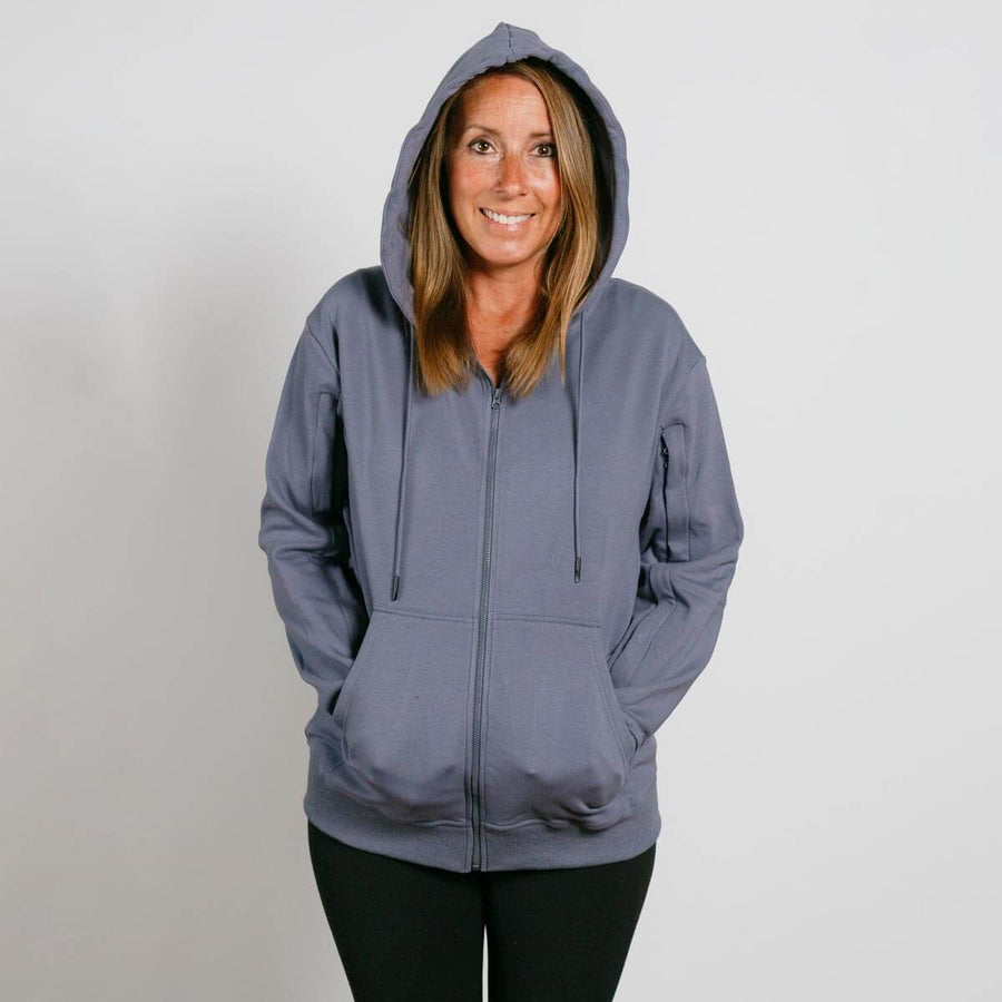 Woman wearing a slate blue zip-up hoodie with hands in pockets, smiling at camera against white background.