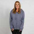 Woman wearing a slate blue zip-up hoodie over black pants, smiling at camera against white background.