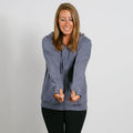 Woman wearing a slate blue zip-up hoodie and black pants, smiling with hands in front pockets.