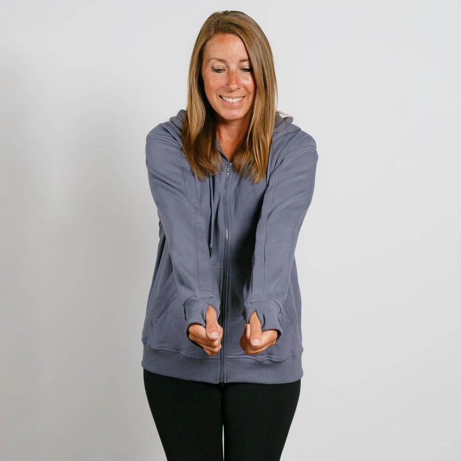 Woman wearing a slate blue zip-up hoodie and black pants, smiling with hands in front pockets.