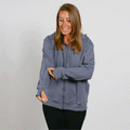 Woman wearing a slate blue zip-up hoodie and black pants smiles while looking down.