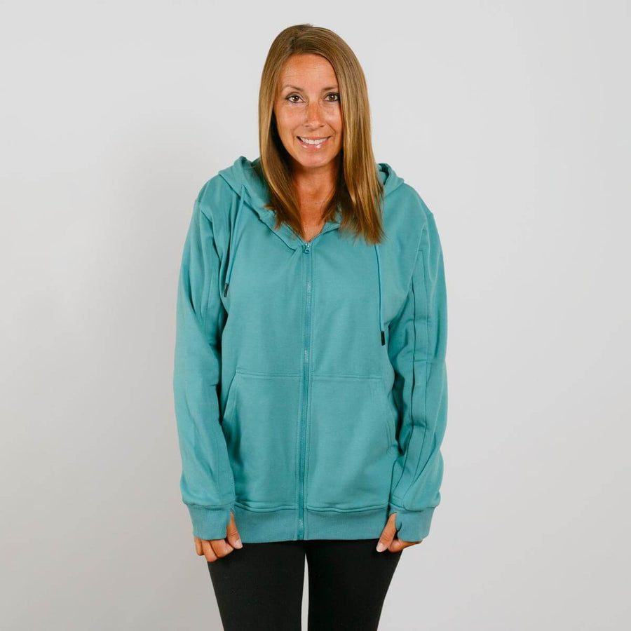 Woman wearing a turquoise zip-up hoodie with black pants against a white background.