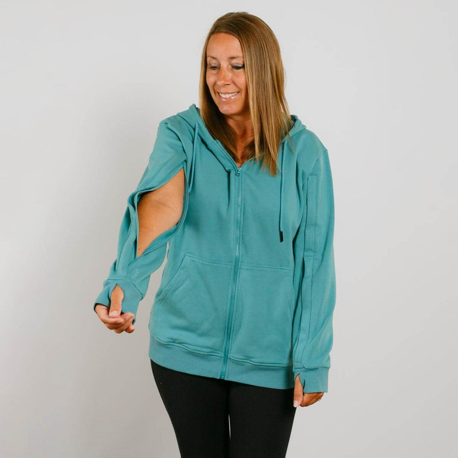 Woman wearing a turquoise zip-up hoodie with cutout sleeves and black pants against white background.