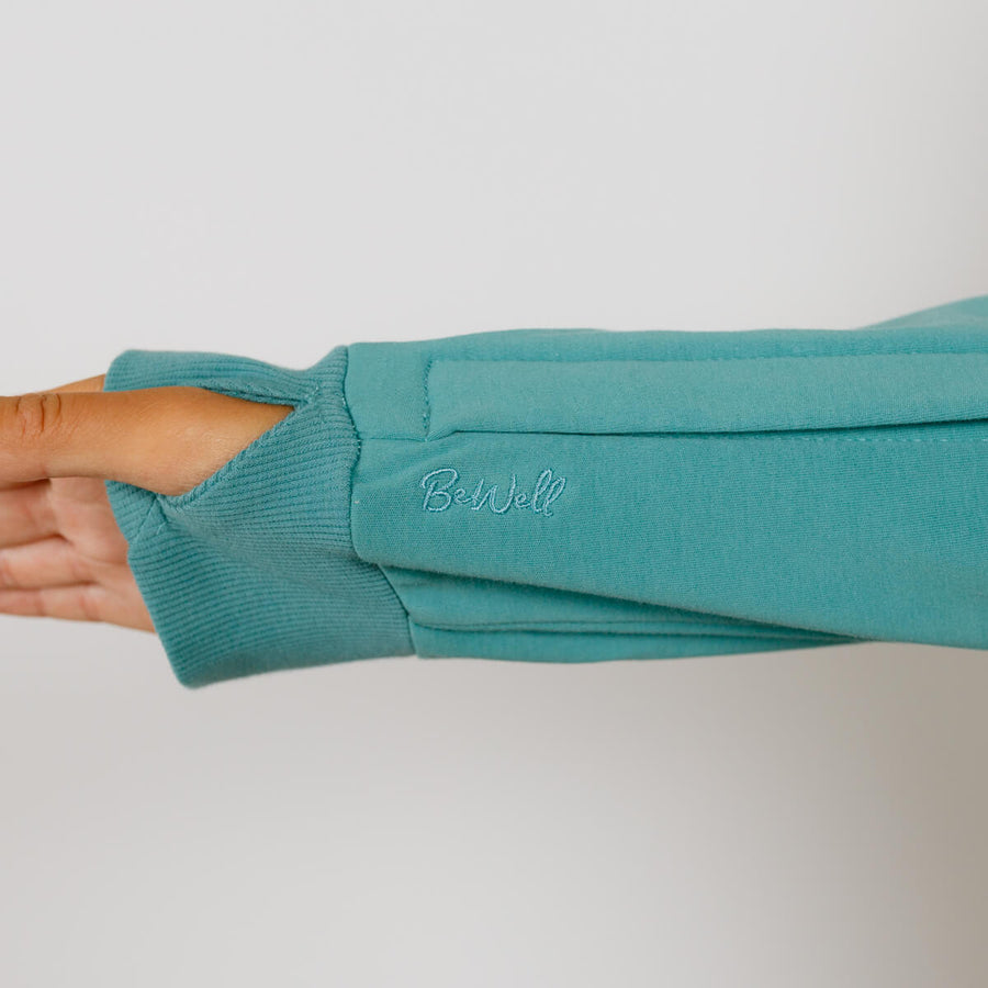 A person's arm extends through a turquoise zip hoodie sleeve against a neutral background.