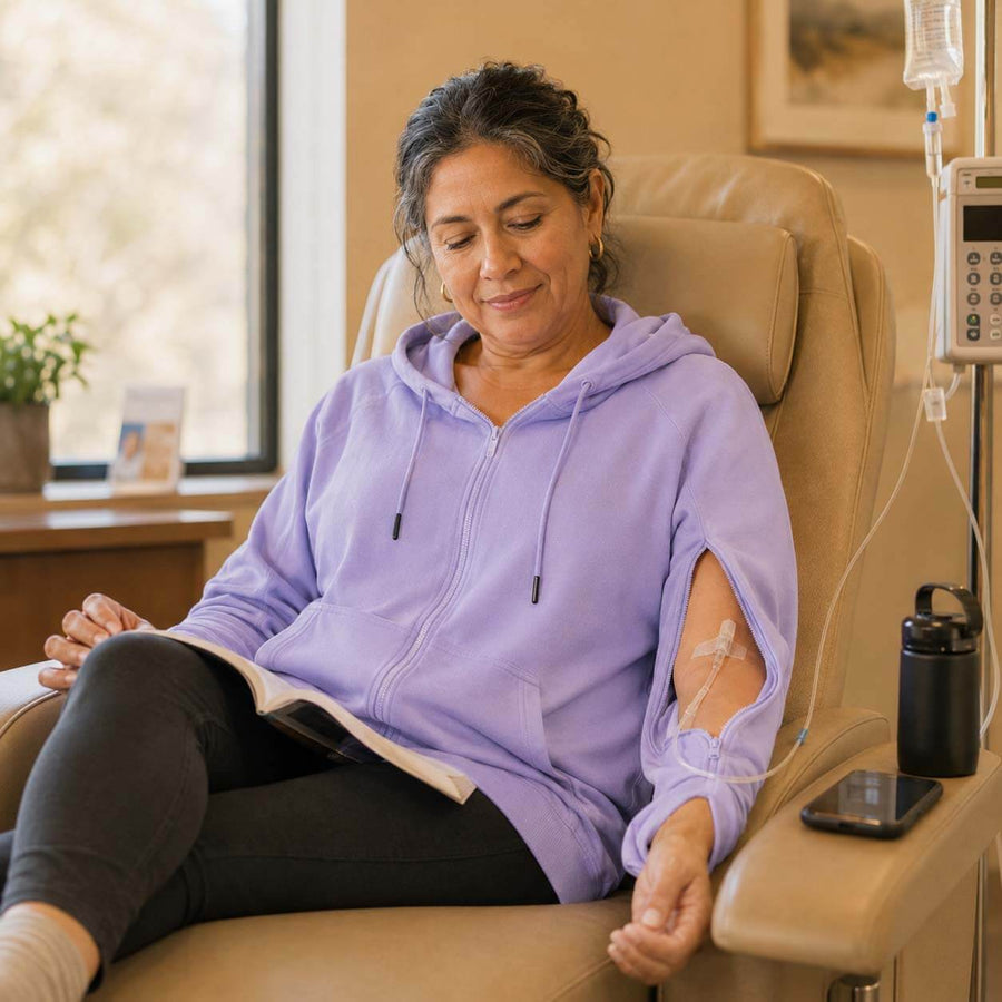 Woman in purple adaptive hoodie with arm openings reads magazine in medical chair with IV stand.