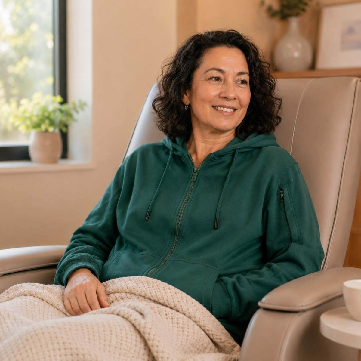 Woman in green hoodie seated comfortably on beige chair in warm, minimalist interior space.