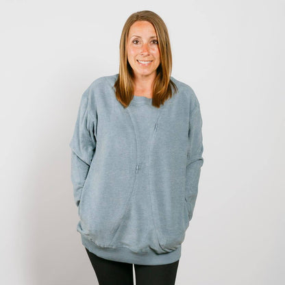 Woman wearing a light blue crewneck sweatshirt with hands in pockets, smiling at camera.