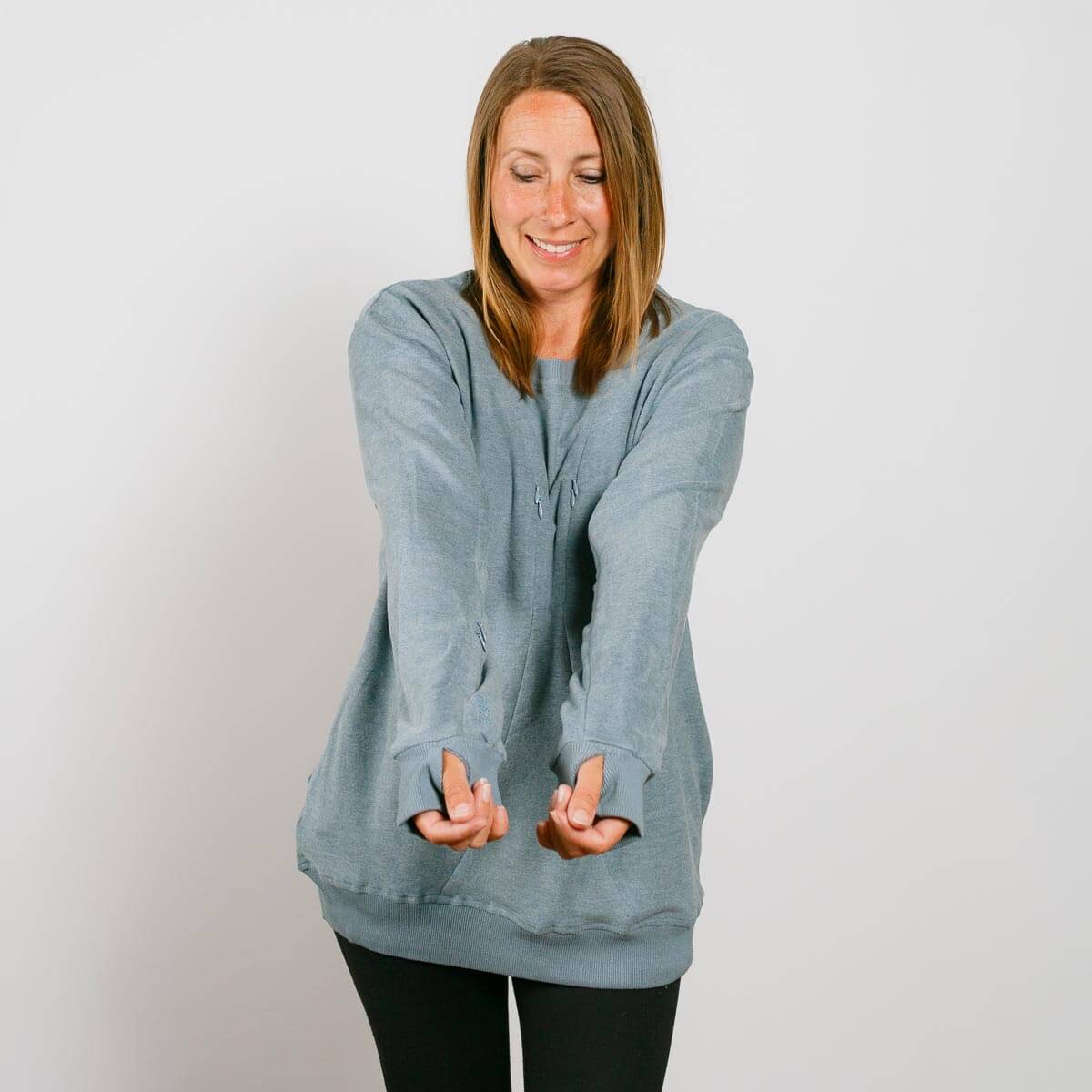 Woman wearing a light blue crewneck sweatshirt with thumb holes, smiling with hands extended downward.