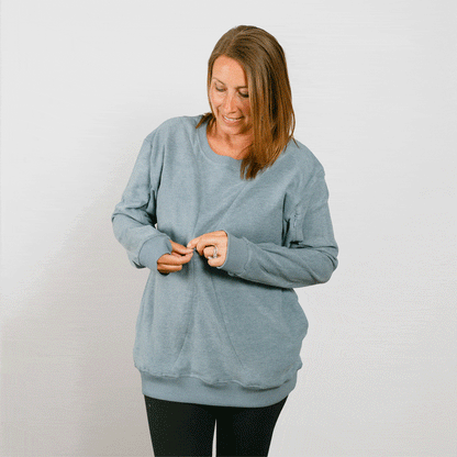 Woman wearing a light blue crewneck sweatshirt over black pants, looking down while adjusting the neckline.