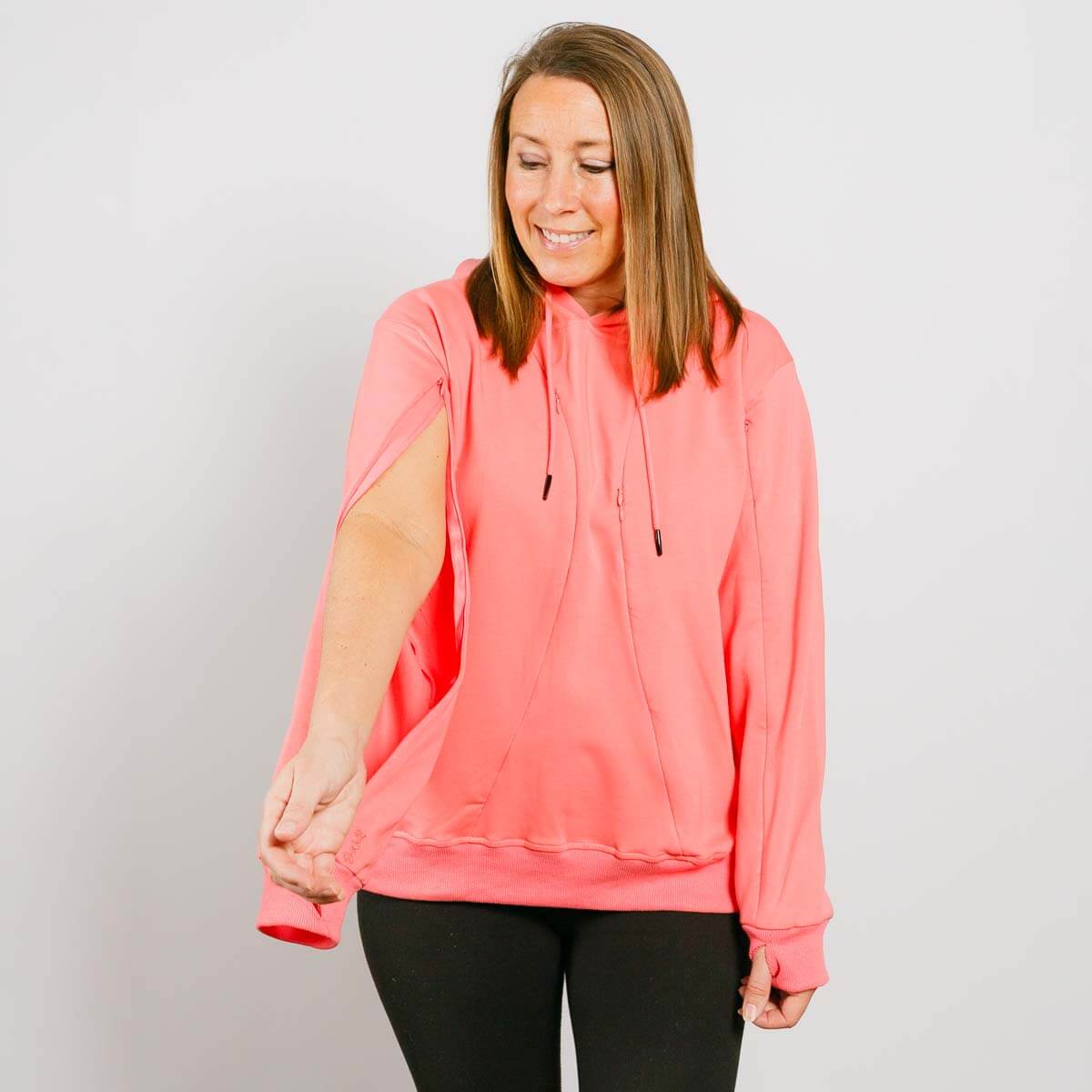 Woman wearing a pink hoodie with black pants, smiling while looking down at her rolled-up sleeve.