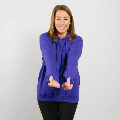 Woman wearing a blue hoodie with black pants, smiling while gesturing with both hands forward.