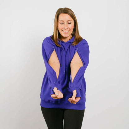 Woman wearing a blue hoodie with open cutouts on the sides, smiling with hands gesturing downward.