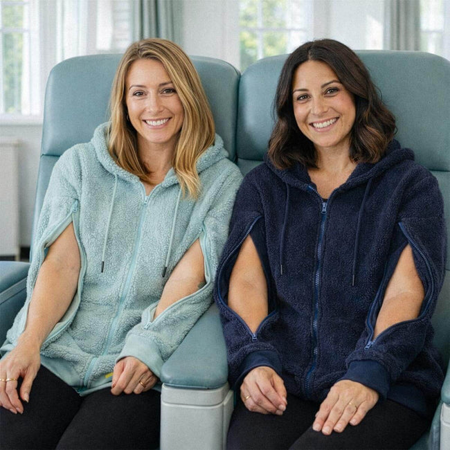 Two women smile while sitting together on a blue chair, wearing adaptive hoodies with open sides.