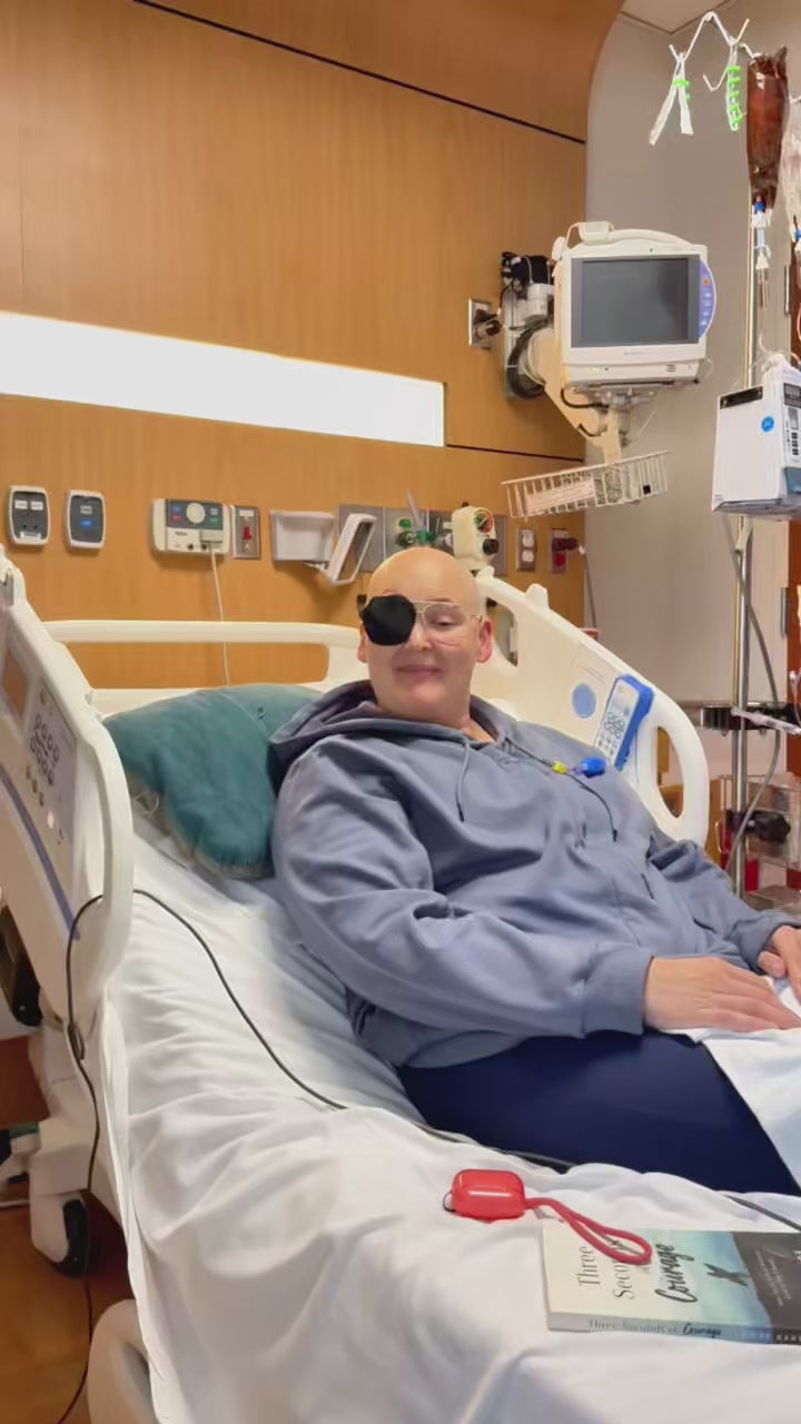 Woman going through chemotherapy, resting in a hospital bed in a blue-gray BeWell hoodie, with an IV pole and monitors behind her.