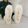 Close-up of white cozy socks with gold "Brave" text on soles, displayed flat.