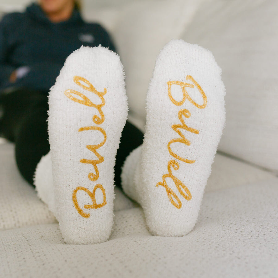 Close-up of white cozy socks with gold "Brave" text on soles, displayed flat.