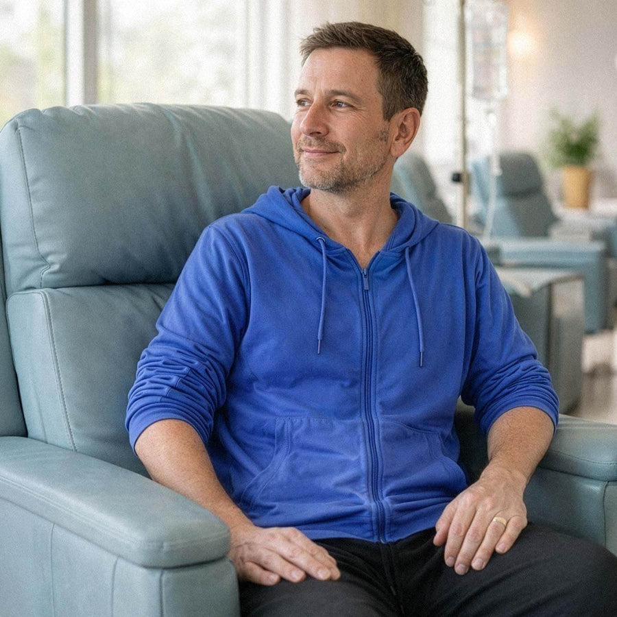 Man in blue zip-up hoodie sitting in gray chair, looking to the side in bright indoor setting.