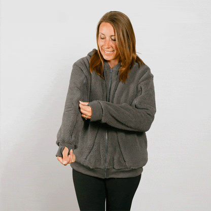 Woman wearing a gray sherpa zip hoodie over black pants, smiling while looking down.