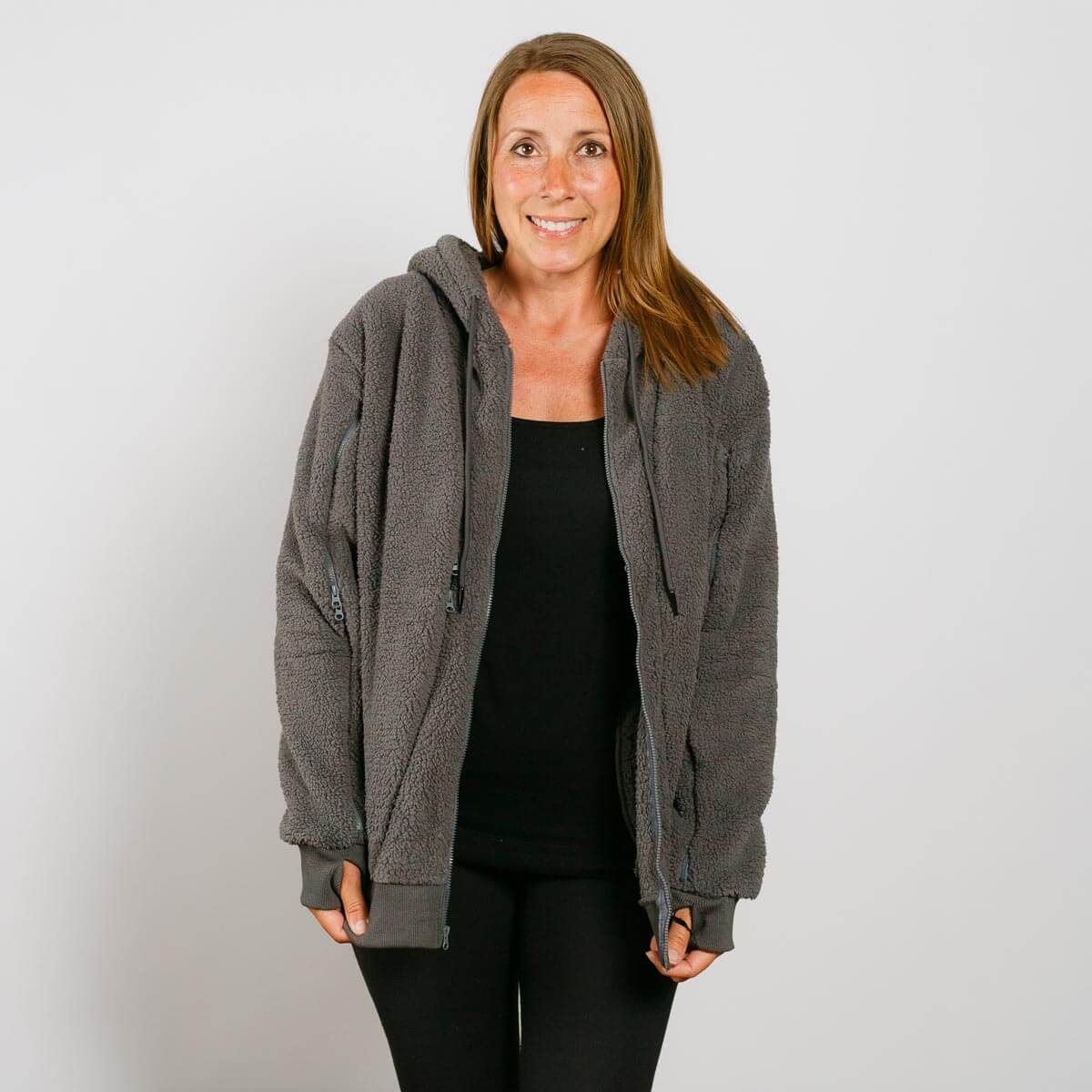 Woman wearing gray sherpa zip-up hoodie over black shirt, smiling at camera against white background.