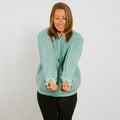 Woman wearing a seafoam color zip hoodie with thumb holes over black pants, smiling while looking down.