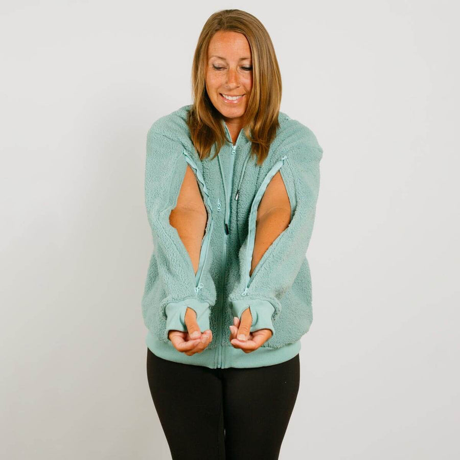Woman wearing a seafoam color zip hoodie with cutout sleeves, smiling while looking down.